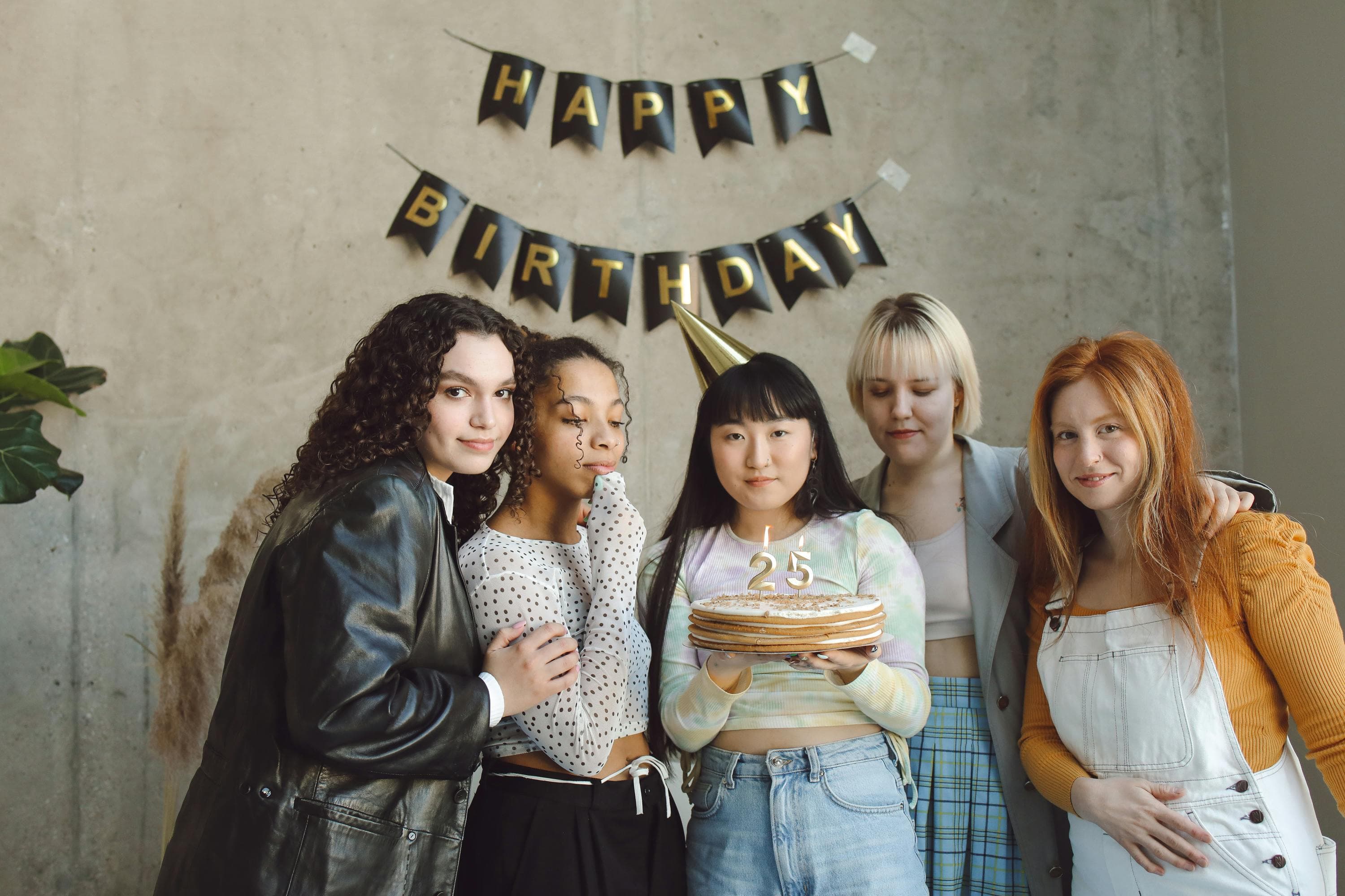 Group of friends at a 25th birthday party holding a cake with candles under a Happy Birthday banner